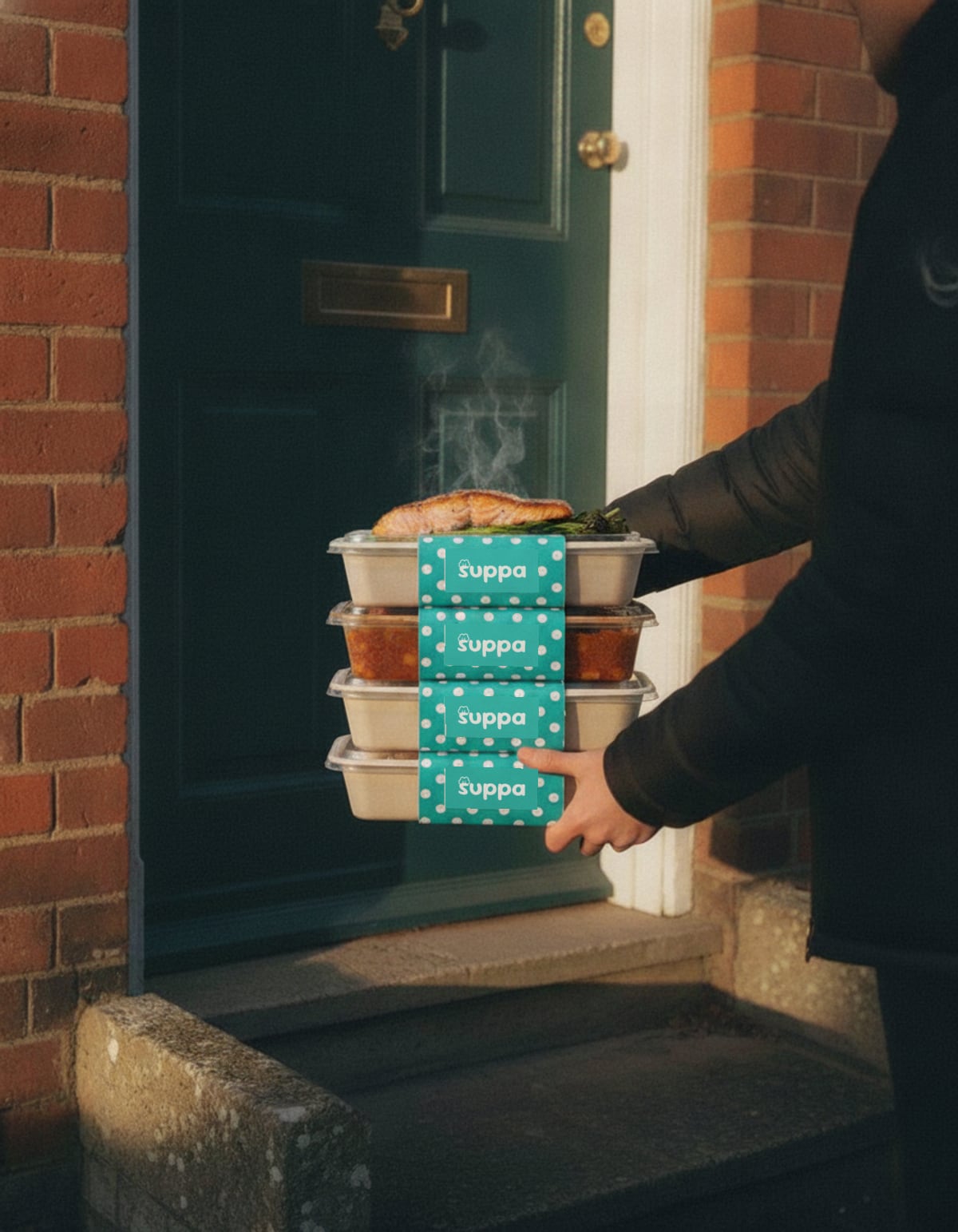 A stack of sleeved Suppa trays being handed over at a front door in the evening